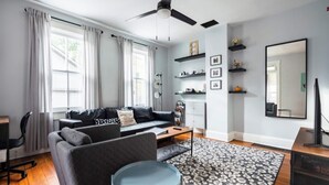 Ein modernes Wohnzimmer mit einer Couch, einem Couchtisch und einer TV-Konsole. Regale mit Dekorationselementen und ein großer Spiegel sind an der Wand angebracht.