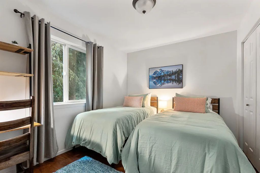 A bedroom with two beds, a window with curtains, a painting on the wall, and a wooden shelf.