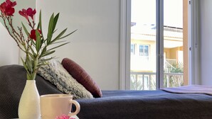 Ein Bett mit dunkelgrauer Bettdecke, eine weiße Vase mit roten Blumen, eine weiße Tasse und eine rosa Blüte auf einem weißen Tisch.