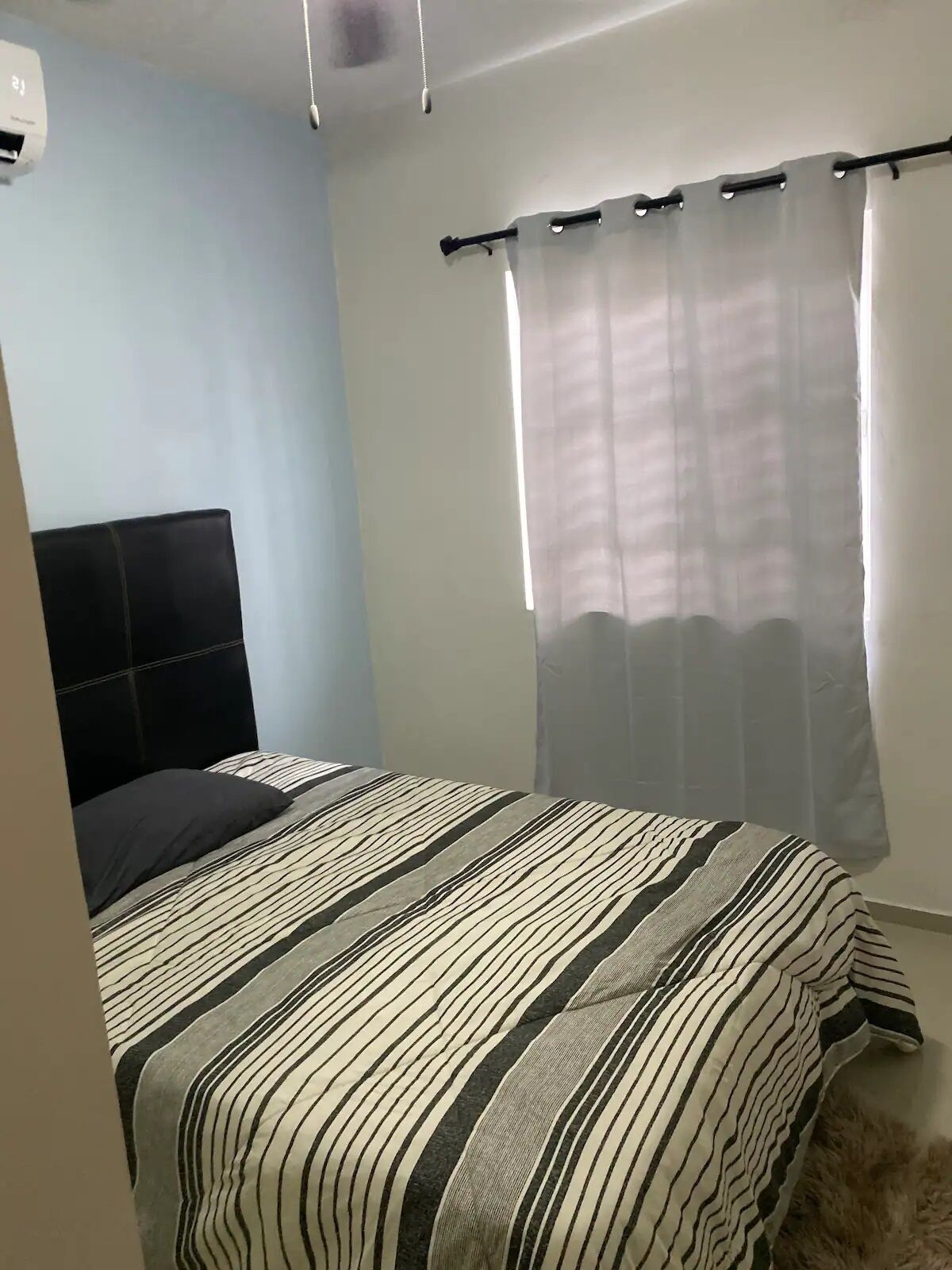 A bedroom with a bed, a headboard, a window with a sheer curtain, and a wall-mounted air conditioning unit.