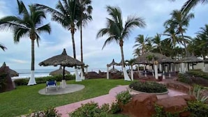 A tropical resort with palm trees, thatched-roof structures, and a pathway leading to a beach. - Departamentos QM2 (Mazatlán)