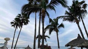A beachside view with tall palm trees, a thatched umbrella, and a chair. - Departamentos QM2 (Mazatlán)