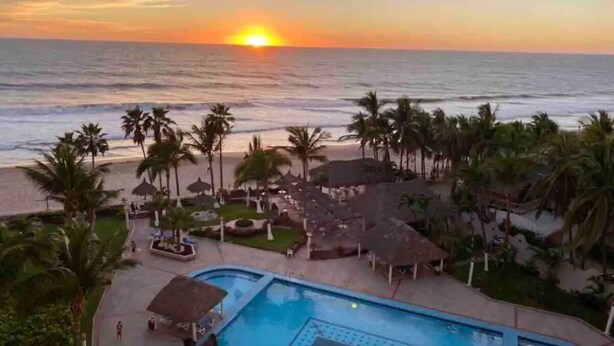 A resort with a swimming pool, palm trees, and a view of the sunset over the ocean. - Departamentos QM2 (Mazatlán)