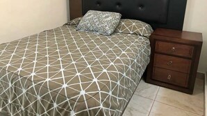 A bed with a geometric patterned bedspread and two pillows, a wooden nightstand with drawers, and a dark headboard. - Departamentos QM2 (Mazatlán)