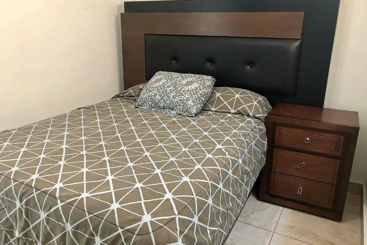 Ein Bett mit geometrisch gemustertem Überwurf und zwei Kissen, ein Nachttisch aus Holz mit Schubladen und ein dunkles Kopfteil.