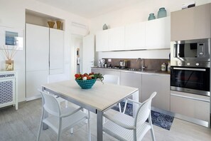 A modern kitchen with white cabinetry, a stainless steel microwave, and an oven. There is a table with chairs and a bowl of fruit.
