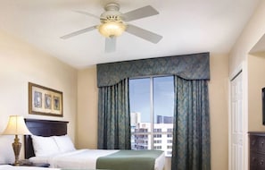 Ein Hotelzimmer mit Bett, Nachttisch, Lampe, Deckenventilator und einem großen Fenster mit Vorhängen.
