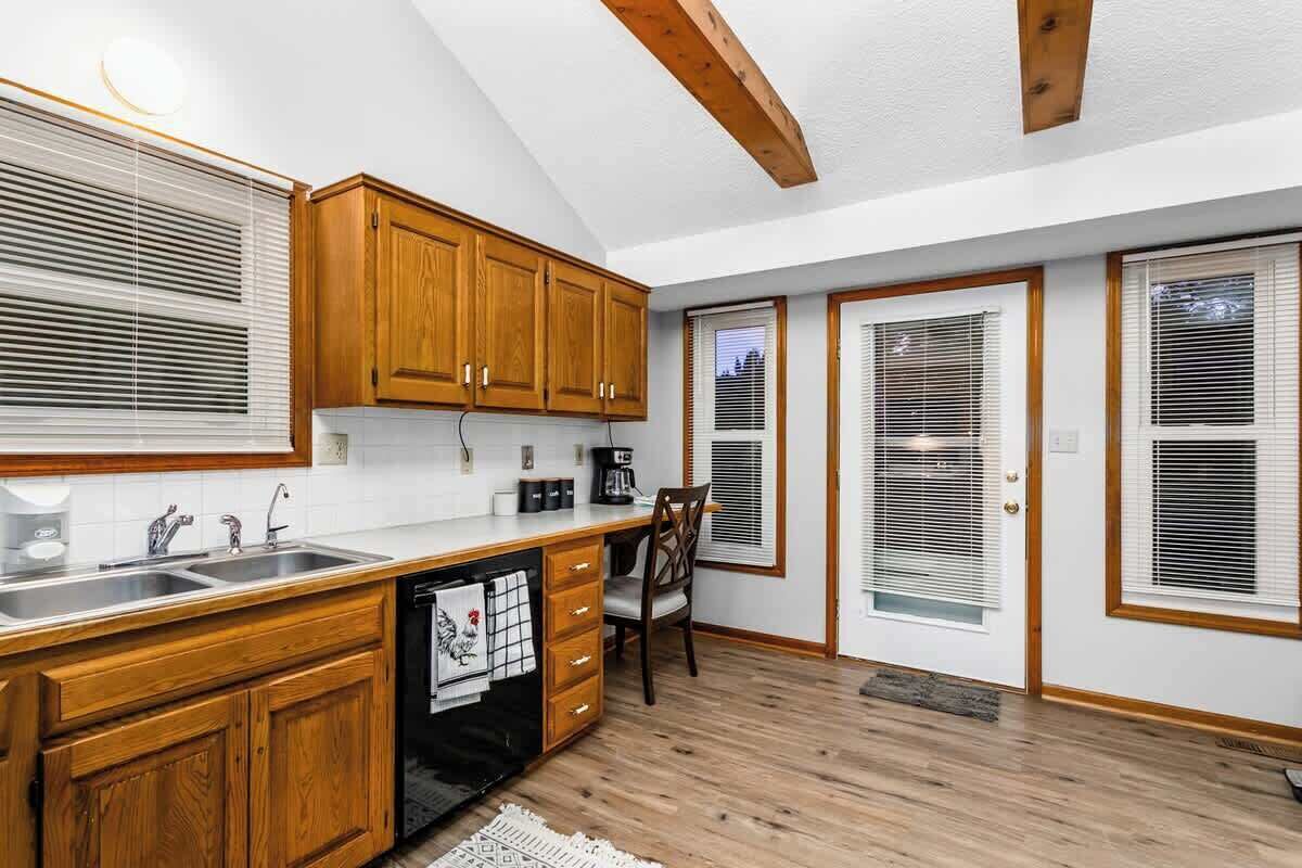 Cocina con armarios de madera, lavavajillas y un lavabo. Hay dos puertas con ventanas y una silla.
