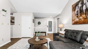 A modern living room with a dark leather sofa, a round wooden coffee table, and a white rug. There is a painting on the wall and a lamp on the side table. - Home Near Legends_Sleeps 8_Great Location_Shops (Kansas City)