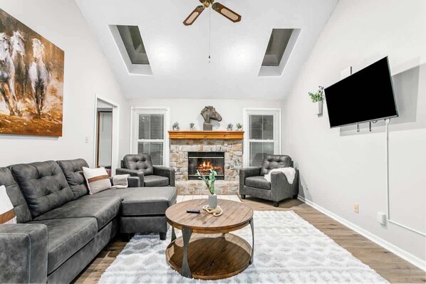 A living room with a sectional sofa, two armchairs, a fireplace, a flat-screen TV, and a ceiling fan.