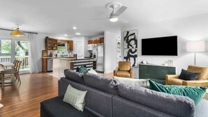 A modern living room with a grey sofa, a coffee table, and a TV. There is a kitchen area with wooden cabinets and a dining table. - Kid-Friendly Home w/ Yard & Workspace (Shawnee)