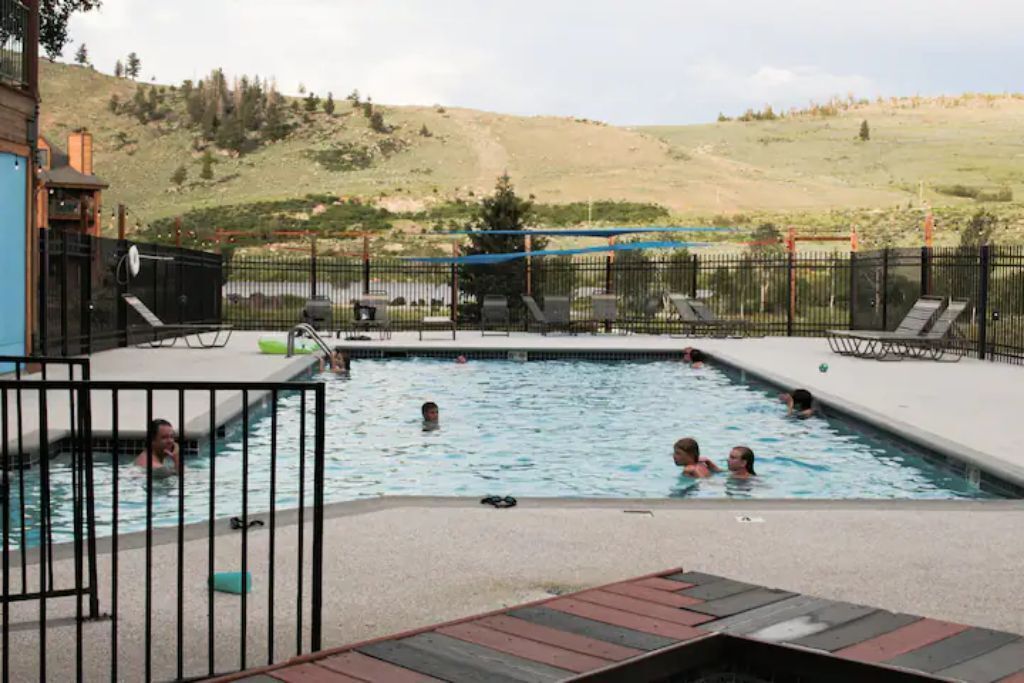 A swimming pool with people in it, surrounded by a fence and a view of a hilly landscape.