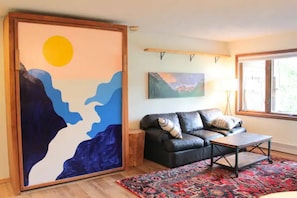 A living room with a black leather sofa, a coffee table, a colorful rug, a painting of a mountain landscape, and a window with a view.