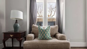 A beige armchair with a patterned pillow, a lamp on a wooden side table, and a window with curtains. - Great Stay_3BR Home_Walk to downtown Lee's Summit (Lee's Summit)