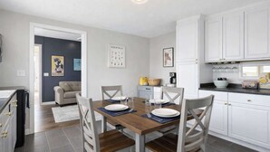 A modern kitchen with white cabinets, a wooden dining table set for four, and a grey sofa in the adjacent living area. - Great Stay_3BR Home_Walk to downtown Lee's Summit (Lee's Summit)