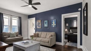 A living room with two sofas, a coffee table, and a ceiling fan. There are framed pictures on the wall and a door leading to another room. - Great Stay_3BR Home_Walk to downtown Lee's Summit (Lee's Summit)