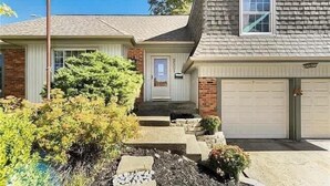 A two-story house with a front garage, a small porch, and a well-maintained garden with various shrubs and a stone pathway leading to the door. - Family Fave w/2 Living Spaces_4BR_Great Location (Overland Park)