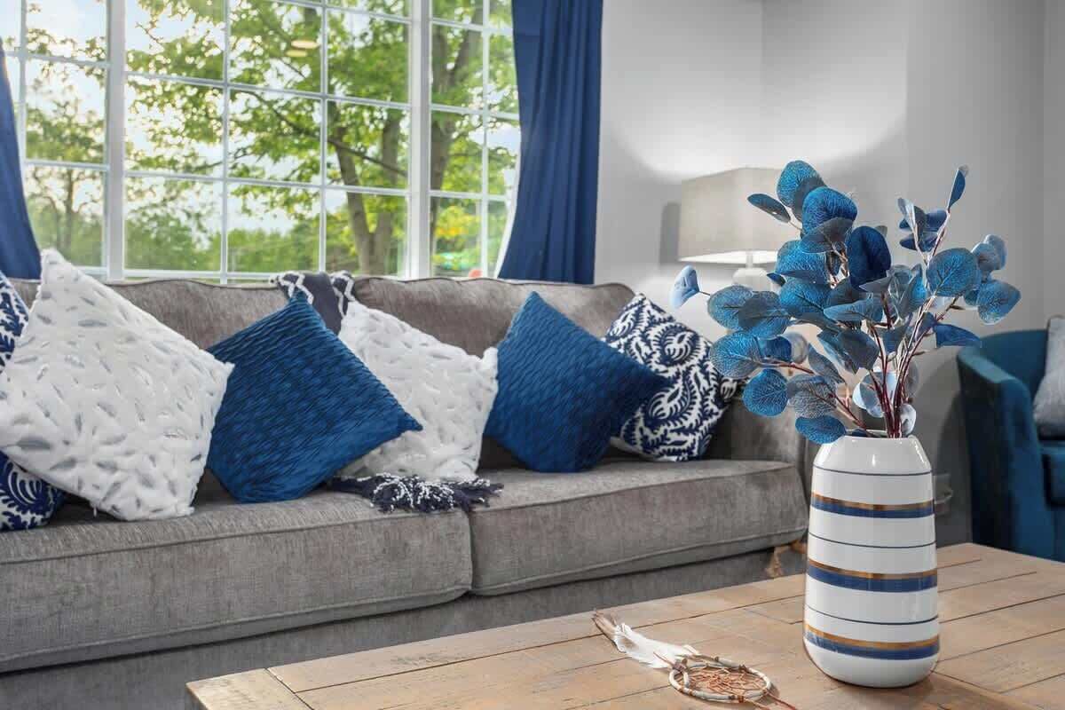 A living room with a grey sofa adorned with blue and white patterned cushions, a white vase with blue and gold stripes on a wooden coffee table, and a view of trees through a large window with blue curtains.