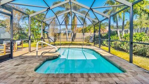 A covered swimming pool with a metal frame, surrounded by a wooden deck and chairs, with a view of palm trees and a fence in the background. - Family Home with Heated Pool_6 Mins from the Beach (Largo)