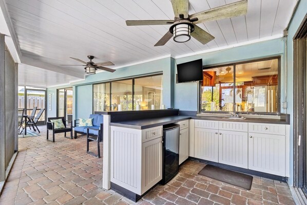 A covered outdoor kitchen area with a built-in refrigerator, a dining table with chairs, and a ceiling fan. - Family Home with Heated Pool_6 Mins from the Beach (Largo)