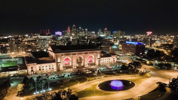 Un edificio grande e histórico con ventanales arqueados y una fuente central, rodeado de una plaza bien iluminada y edificios modernos al fondo.