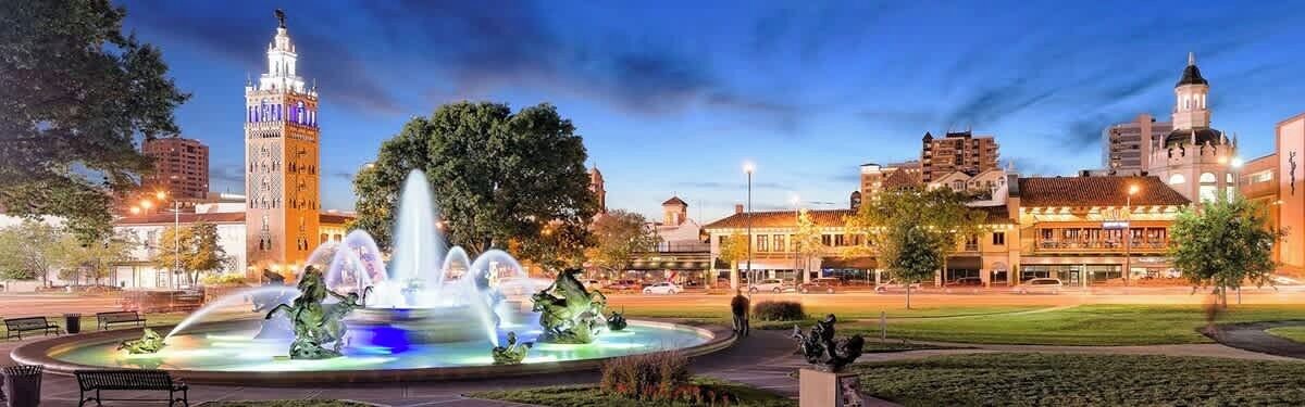 A city square with a fountain, a clock tower, and buildings in the background.
