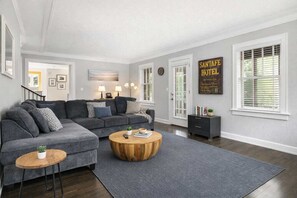 A living room with a grey sectional sofa, a round wooden coffee table, and a grey rug. There is a black side table with books and a plant. The room has white walls, a large window with blinds, and a door leading to another room.