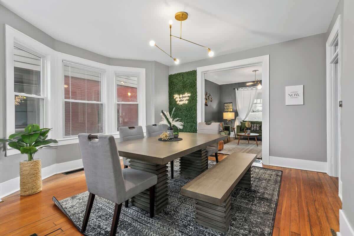 Uma área de refeições com uma mesa grande, cadeiras e um banco. Há uma parede verde com uma luminária decorativa, e uma janela com persianas.