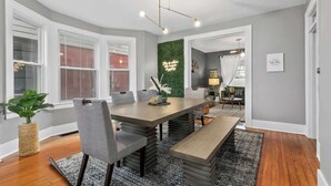 Uma área de refeições com uma mesa grande, cadeiras e um banco. Há uma parede verde com uma luminária decorativa, e uma janela com persianas.