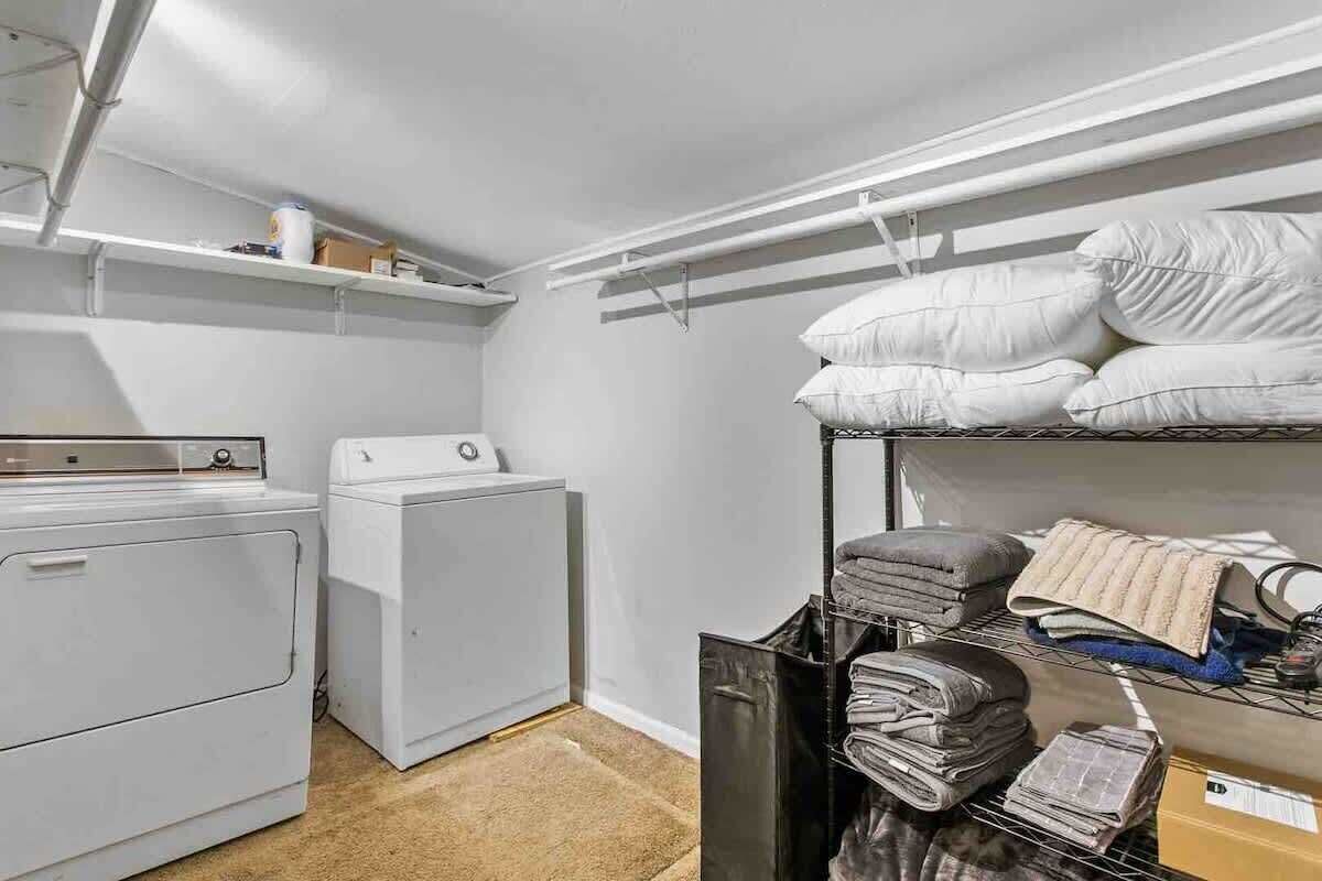 Un lavadero con dos lavadoras blancas, un estante con productos de limpieza y una cama litera con almohadas y mantas dobladas.