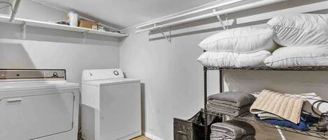 Un lavadero con dos lavadoras blancas, un estante con productos de limpieza y una cama litera con almohadas y mantas dobladas.