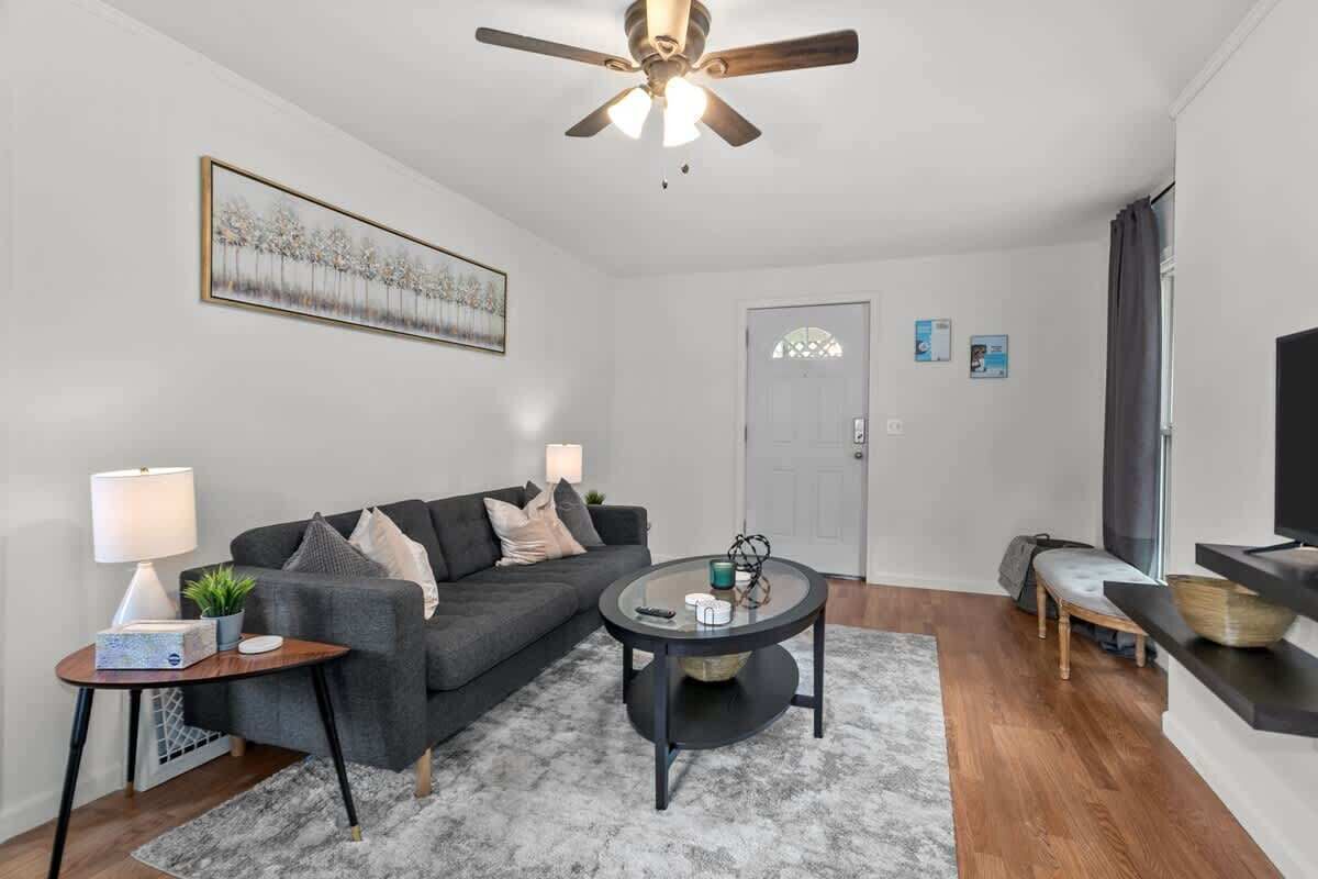A modern living room with a grey sofa, a round coffee table, a ceiling fan, a TV on a stand, and a door with a window.