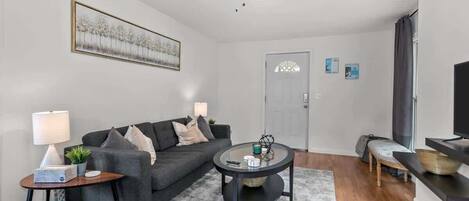 Una sala moderna con un sofá gris, una mesa de centro redonda, un ventilador de techo, un televisor sobre un mueble bajo y una puerta con un ventanal.