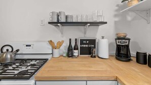 Una cocina con estufa a gas, cafetera y repisa con tazas y un cartel.