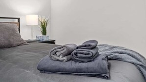 Una cama bien hecha con una manta gris y toallas enrolladas, una lámpara de noche y una pequeña planta en una mesita.