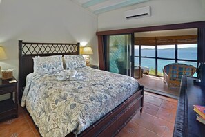 A bedroom with a bed, a nightstand, a lamp, and a view of the ocean through large windows.