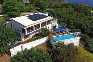 A modern two-story house with a rooftop solar panel, a swimming pool, and a deck with lounge chairs.