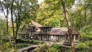 Un chalet rustique aux fondations en pierre, entouré d’une forêt dense. Il dispose d’une grande terrasse avec des meubles d’extérieur et d’un chemin en pierre menant à l’entrée.