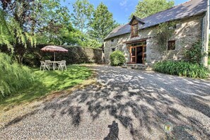 Un bâtiment en pierre avec une allée de gravier, une table et des chaises en bois sous un parasol rouge, et un mur de pierre recouvert de plantes grimpantes.