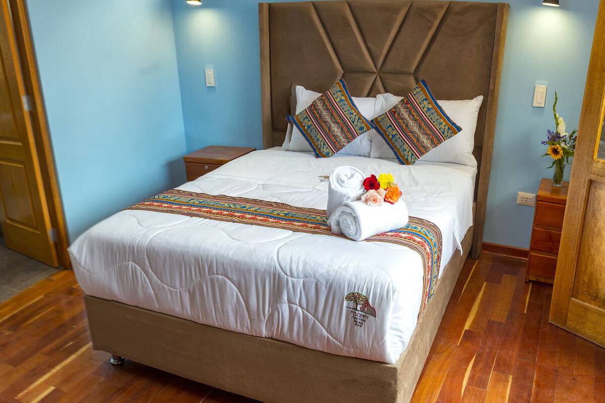 Una cama bien hecha con una colcha estampada, dos almohadas y una toalla enrollada con flores.