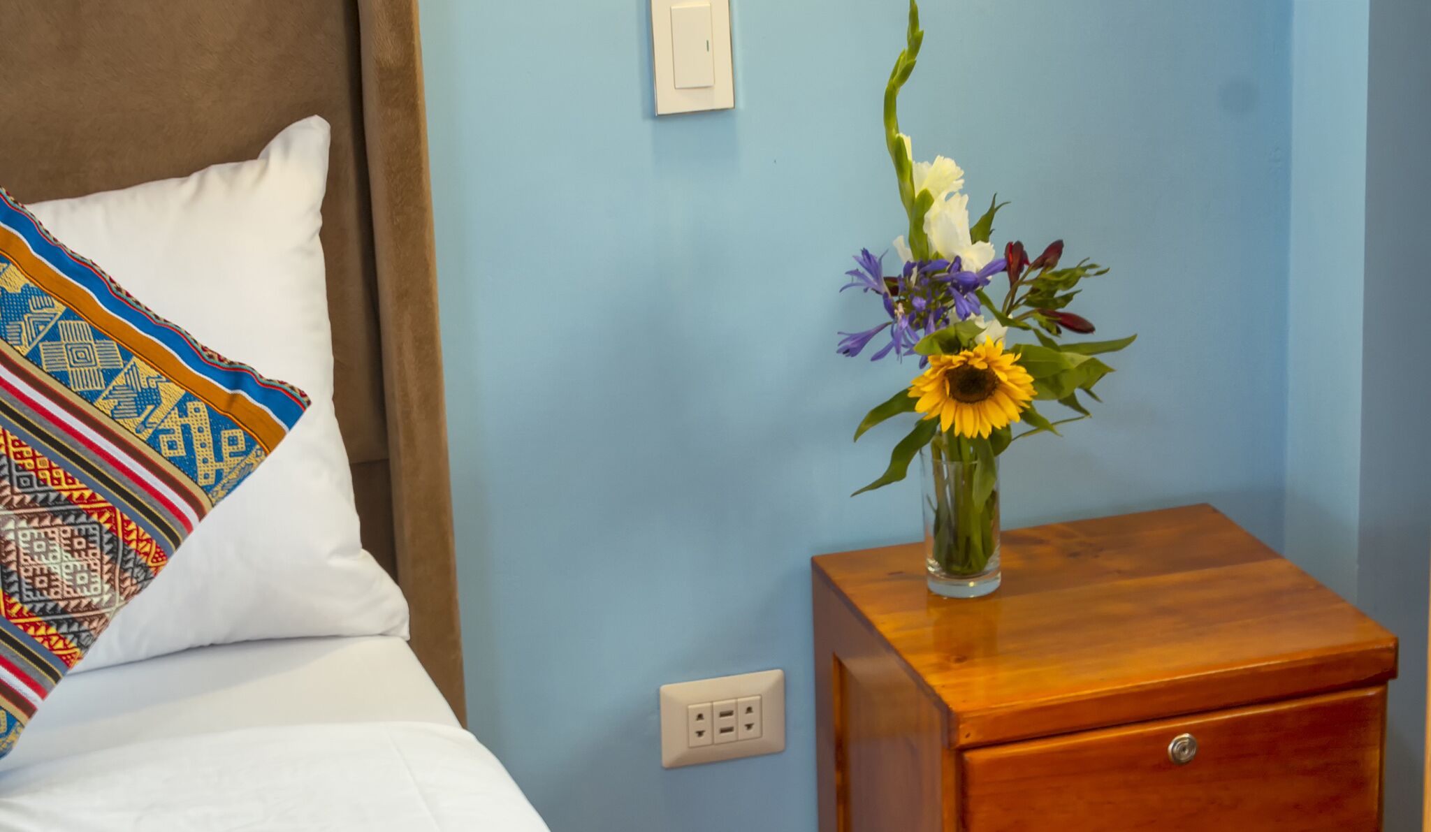 Un lit avec un oreiller blanc et une couverture colorée, une table de chevet en bois avec un vase de fleurs, et un mur bleu avec un interrupteur.
