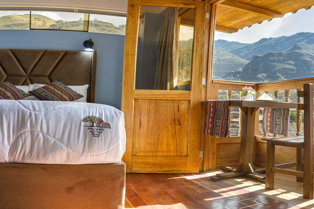 Un dormitorio con cama, cabecera de madera, mesita de noche, ventana con vistas a montañas y balcón con barandilla de madera.