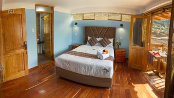 A large bed with a headboard, wooden floors, a bathroom with a shower, and a balcony with a view.