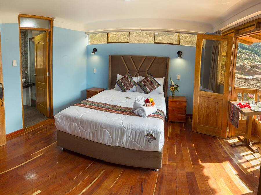 A large bed with a headboard, wooden floors, a bathroom with a shower, and a balcony with a view.