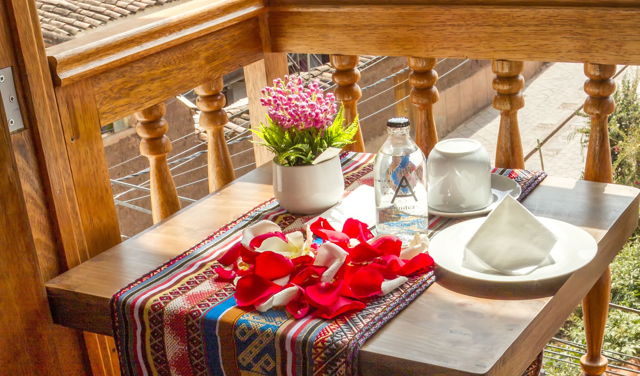 Un balcón con barandilla de madera, una mesa con mantel colorido, una planta en maceta, una botella, una taza y un plato.