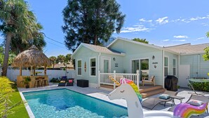 Una piscina con un giostra a forma di unicorno, un solarium con mobili da esterno e una casa con recinzione bianca e patio coperto.