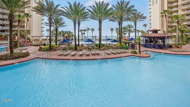 A pool area with palm trees, lounge chairs, and a small building, surrounded by high-rise buildings. - Beachfront Paradise with Reserved Parking (Panama City Beach)