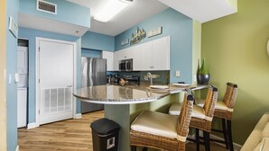 A kitchen with a granite countertop, stainless steel appliances, wicker chairs, and a trash can. - Beachfront Paradise with Reserved Parking (Panama City Beach)
