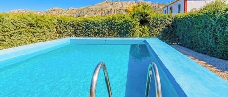 Une piscine d’un bleu éclatant, dotée d’échelles en métal, entourée de verdure et bordée d’un bâtiment au toit rouge.