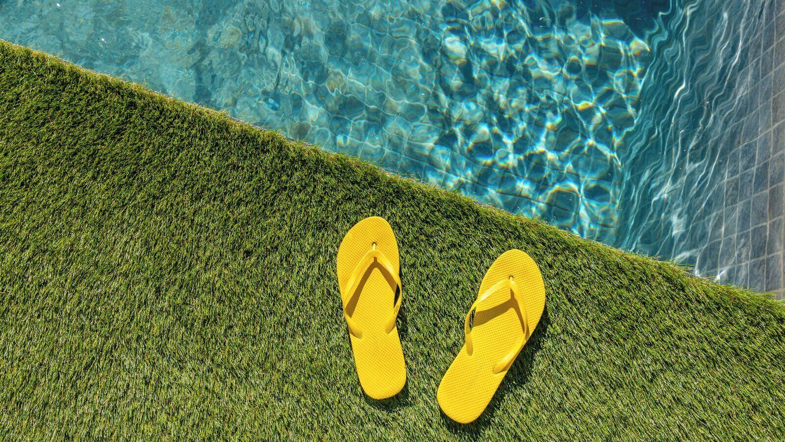 A pair of yellow flip-flops on a green artificial grass surface next to a clear blue pool.